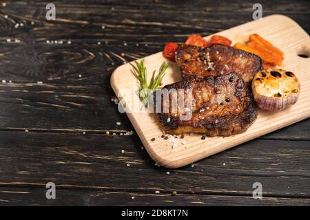 A juicy piece of fried meat with grilled cherry tomatoes garlic and carrot lies on a cutting board against a black wooden table. Degree of roasting we Stock Photo