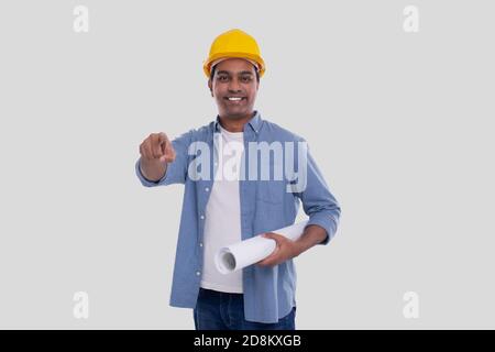Construction Worker Holding House Plan in Hands Pointing in Camera. Architect Holding Blueprints. Yellow Hard Helmet. Stock Photo