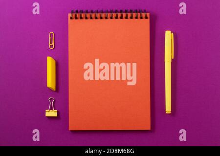 An orange notebook on a bright purple background with yellow office supplies on it, a pen and an eraser and a clamp and a clip. Stock Photo