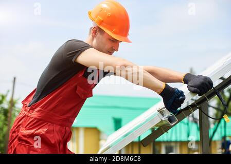 Young worker installing solar panels near the house. A man is wearing an orange uniform, hard hat, black t-shirt and gloves on a warm day. Alternative energy sustainable concept Stock Photo