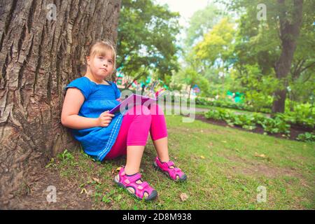 A portrait of trisomie 21 child girl outside having fun on a park reading book Stock Photo