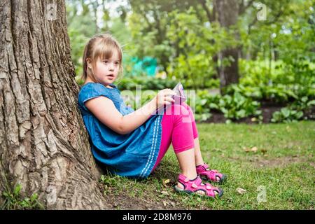 A portrait of trisomie 21 child girl outside having fun on a park reading book Stock Photo