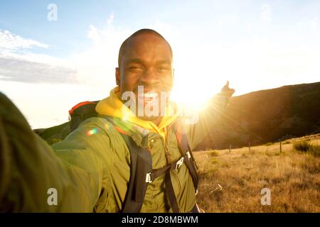 Portrait of happy young black man hiking with backpack taking selfie and pointing to sunset Stock Photo