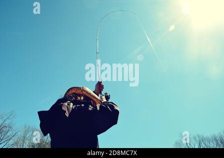 Man wearing a cowboy hat, with back turned to the camera. Holding a bent fishing pole up high, with line catching the glare of the sun. Stock Photo
