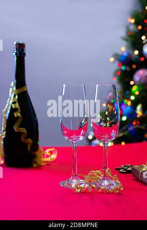 A bottle of champagne and two glasses on a red table with a Christmas tree in the background. New Year 2021, vertical photo. Stock Photo