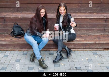 Two young brunette women sit in park in autumn on a wooden bench and take a selfie on the phone in black jackets and smile and hold coffee cups in the Stock Photo