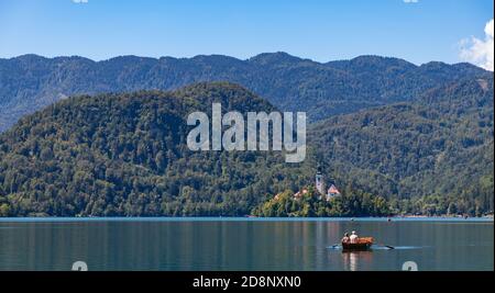 A picture of the Lake Bled Island, the surrounding landscape, and people rowing a boat. Stock Photo