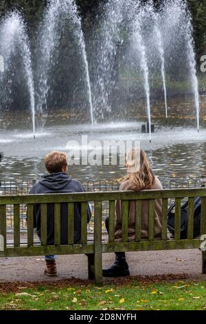 a young couple of lovers sitting on a wooden bench looking at a fountain in the central park in leamington spa, warwickshire, uk. Stock Photo