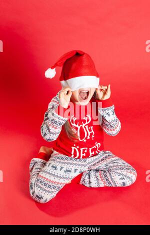 a happy girl in red Christmas pajamas with reindeer hides under a Santa Claus hat on a red background Stock Photo