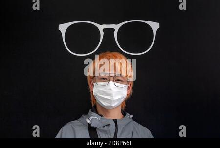 A male, Asian youth, wearing a face mask and spectacles, poses in front of a spectacle sign in London’s Soho district. England, UK Stock Photo