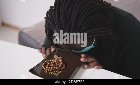 Black African Descent Man With Face Mask Praying on Holy Bible Book and Rosary. Seeking Hope in Religion During Covid-19 Virus Pandemic Outbreak. High quality photo