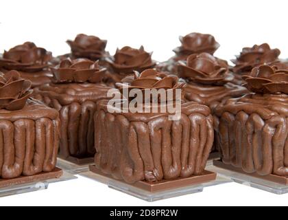 Square chocolate cake cupcakes on a chocolate candy bar frosted with choc frosting, handmade chocolate  rose on top, sitting on square clear plates is Stock Photo