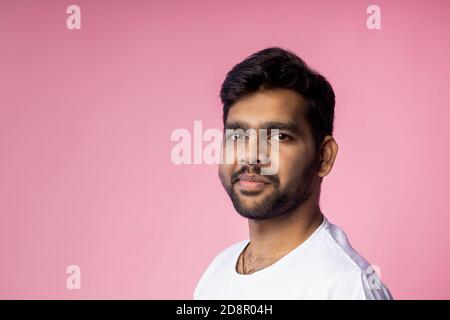 portrait of handsome young indian man with stubble, wearing white t shirt, looking at camera with serious, calm expression, isolated over pink backgro Stock Photo