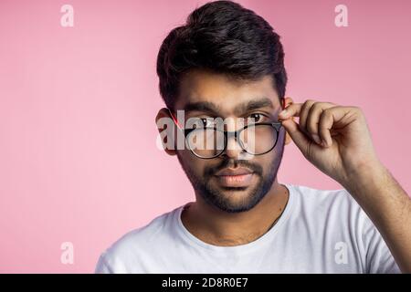 Handsome smart indian guy, student dressed in white t shirt, smiling looking at camera, wearing glasses and touching it, posing in studio over pink ba Stock Photo