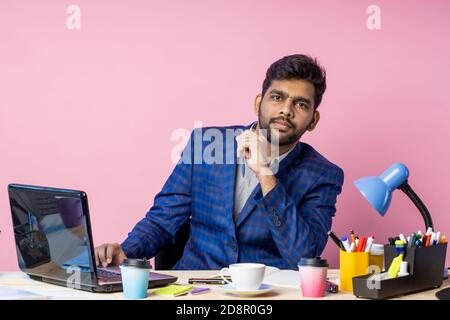 Portrait of tired pensive young indian entrepreneur sitting at workplace, holding pen, thinking about financial problems, wearing suit, looking though Stock Photo