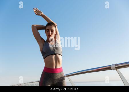 Image of young attractive woman stretching body before running workout at the seaside promenade. Sportswoman warm-up for jogging and training on pier
