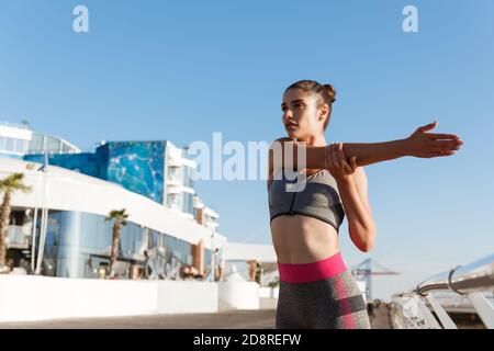 Young attractive woman stretching hands and warming-up before workout. Runner doing exercises on the seaside pier, preparing for jogging Stock Photo