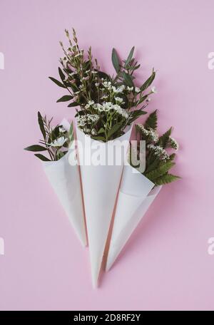top view closeup of three beautiful bouquets of delicate small white flowers with green leaves in paus tracing paper cone on pink background copyspace Stock Photo