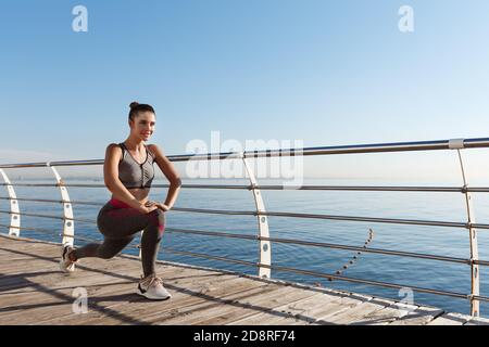 Outdoor shot of smiling fitness woman stretching and working out on the seaside promenade