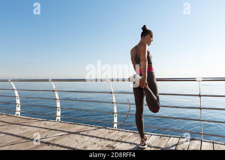 Rear view of attractive fitness woman stretching her legs before jogging, leaning on handrail along seaside promenade, training outdoors Stock Photo