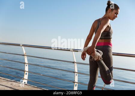 Rear view of sportswoman stretching her leg before running along seaside Stock Photo