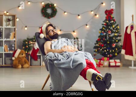 Tired young man dressed as Santa Claus sleeping in an armchair under warm blanket Stock Photo