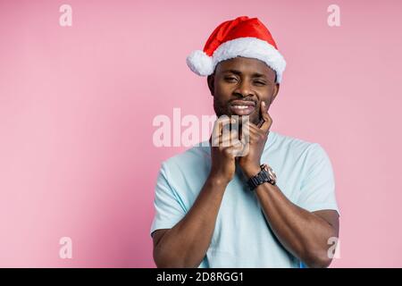 Thoughtful dark skinned man with stubble in Santa hat keeping hands on face, trying to make decision, or remember something important, thinking isolat Stock Photo