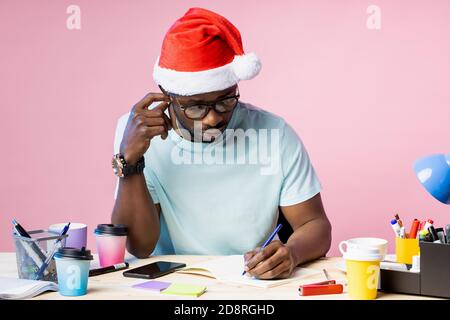Serious young african american male designer in eyewear, dressed in white t shirt, Santa Hat, sitting at table writing new ideas in notepad, posing on Stock Photo