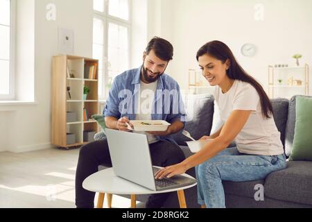 Young man and woman sitting on sofa in the day and eating takeaway food and using laptop. Stock Photo