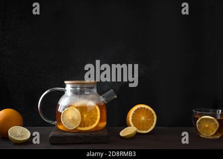 Citrus tea with  lemon, oranges  in transparent glass teapot on dark wooden table, copy space. Fruit tea. Seasonal winter autumn hot drink. Stock Photo