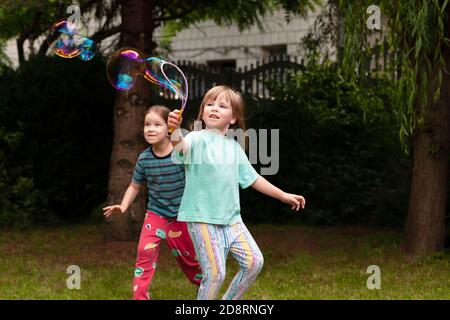 Two happy little girls running around playing with soap bubbles in the backyard Active healthy young children playing and making bubbles in the garden Stock Photo