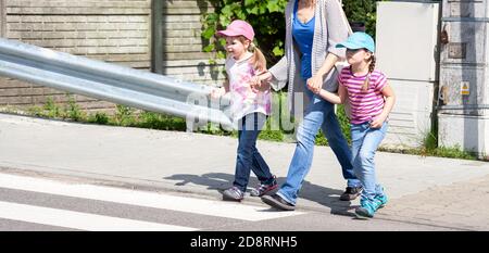 Two little girls, children crossing the street on the zebra crossing holding hands with their mother. Happy sisters safely going across the street Stock Photo