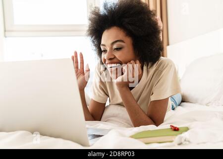 Cheerful young african woman having video chat with friends using laptop camera while lying on bed, waving hand