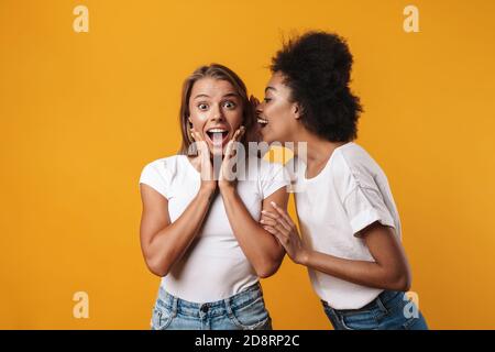 Photo of emotional happy young multiracial girls friends gossiping isolated over yellow wall background Stock Photo