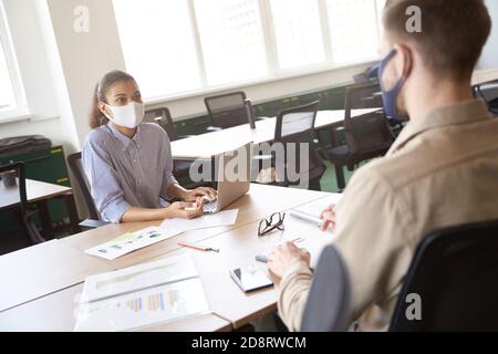 Young mixed race woman wearing protective face mask using laptop and discussing something with male colleague while working together in office during Stock Photo