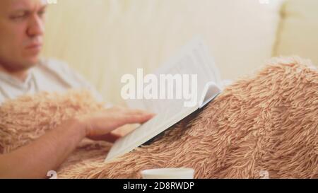 Man reading a book while sitting on the sofa under the covers, blurred background Stock Photo