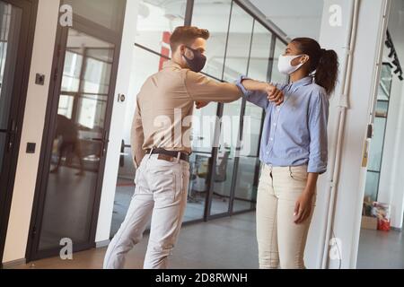 Two young diverse colleagues wearing face protective masks bumping elbows, greeting each other while standing in the modern office Stock Photo