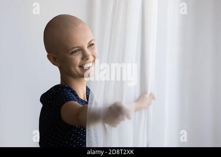 Close up smiling dreamy hairless woman opening curtains in morning Stock Photo