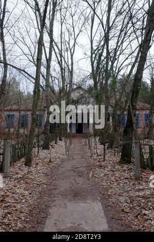 The facade of an abandoned kindergarten in the Chernobyl radioactive zone. Autumn foliage on the ground. Trees in front of an old abandoned house in t Stock Photo