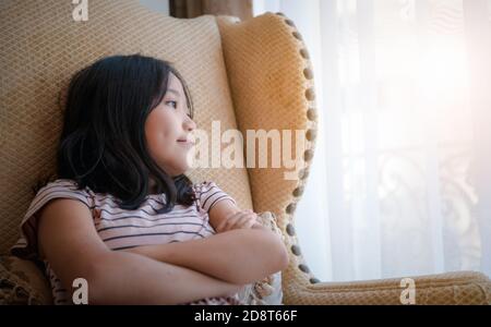 Beautiful little asian girl smiling and watching out the window. A child looks out the window. Young girl looking from window. Stock Photo