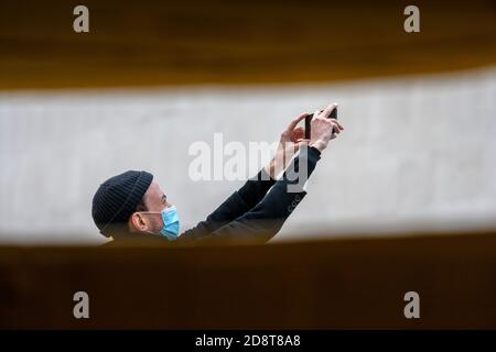 Man, wearing a beanie hat and facial mask, using his mobile phone to make a digital video. London’s Southbank, UK Stock Photo
