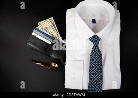 folded white dress shirt with necktie and money in black leather wallet and pipe on black background Stock Photo
