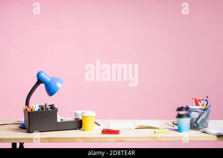 Workplace in office. Stationery, notepad, paper cups, table lamp, on desktop. Office supplies. Colorful workspace. Pink background with copy space for Stock Photo