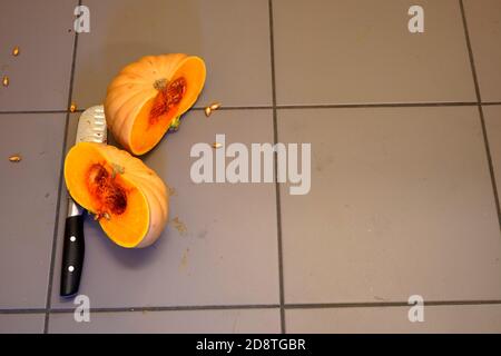 A butternut squash or pumpkin cut into two halves with a knife fallen on the ground in a kitchen. The seasonal vegetable in bright orange color. Stock Photo