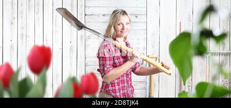 smiling woman with spade in flower garden isolated on white wooden shed background with gardening tools and copy space Stock Photo