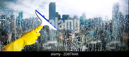 Cleaning windows with skyscraper on background. Hand in yellow glove hold cleaning squeegee. Cleaning service concept. Stock Photo