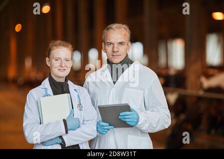 Waist up portrait of two smiling veterinarians at farm looking at camera while holding tablets, copy space Stock Photo