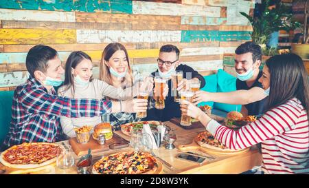 Happy young friends with protective face masks eating and drinking beer at restaurant Stock Photo