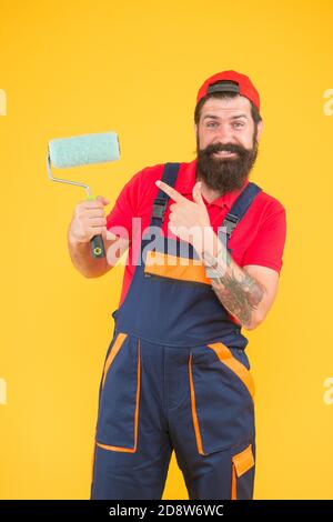 smiling bearded guy painter worker in boiler suit uniform pointing finger on paint roller tool for facing wall, repair. Stock Photo