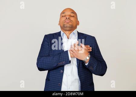Portrait of grateful young African American businessman guy smiling with hands on chest with closed eyes and grateful gesture on face. Health concept. Standing on a gray background. Stock Photo